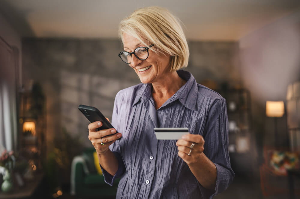Imagem de uma senhora idosa segurando um cartão benefício consignado em uma mão e o celular em outra.