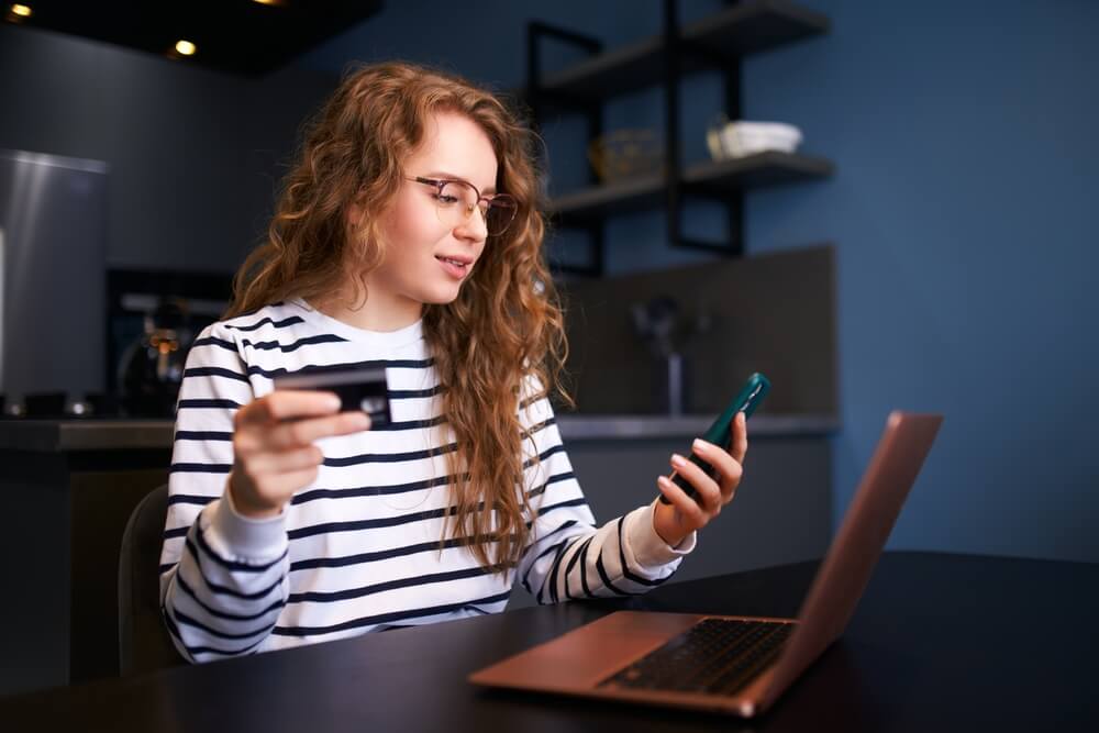 Imagem de um menina ruiva com óculos. Ela está segurando um cartão em uma mão e o celular em outra. Além disso, há um computador na frente dela na mesa.