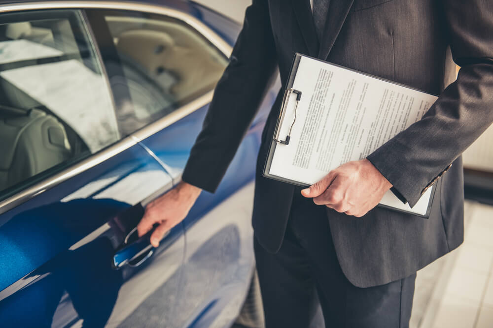 Imagem de um homem segurando um contrato e abrindo a porta de um carro azul. Ele está de terno cinza.