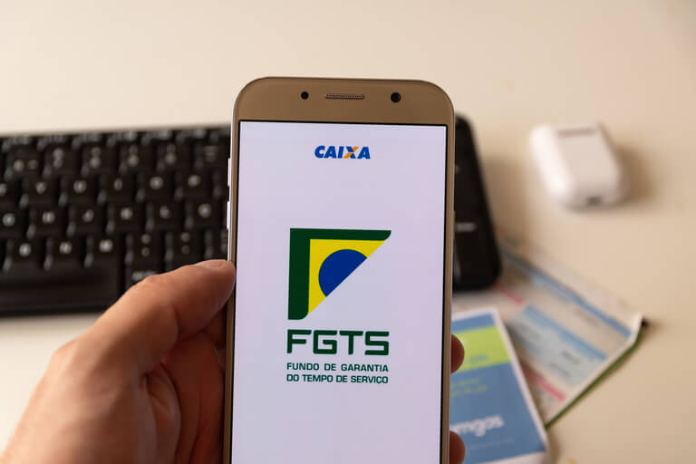 Imagem de uma mão segurando um celular. A pessoa está abrindo o aplicativo do FGTS. Ao fundo, é possível ver um teclado de computador, além de contas de energia e água.