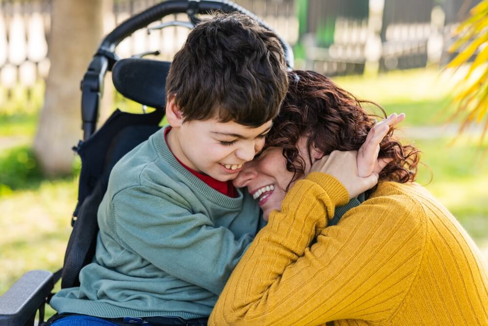 Imagem de uma criança com deficiência abraçando uma mulher. Aparentemente, eles estão em um parque.