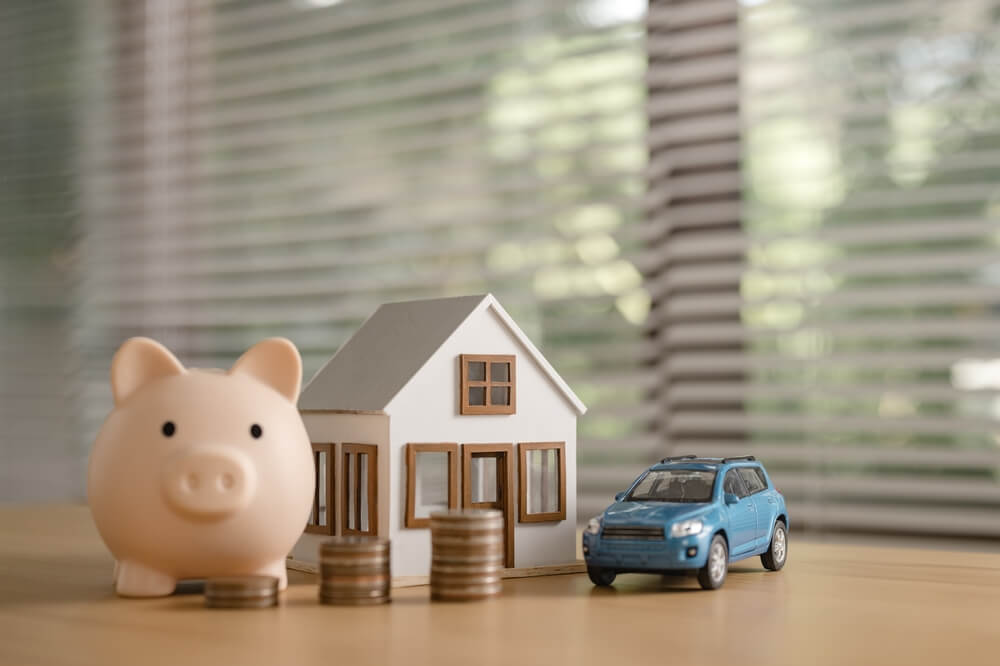 Imagem de um porquinho, moedas, uma casa e um carro azul em cima de uma mesa.