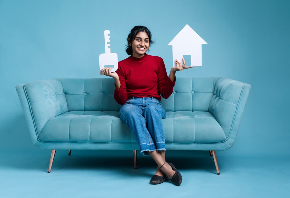 Imagem de uma mulher morena sentada em um sofá azul. Ao fundo, é possível notar uma parede no mesmo tom que o sofá. Ela está segurando uma chave em uma mão e, na outra, um casa. Ambos de papel, representando o Home Equity.