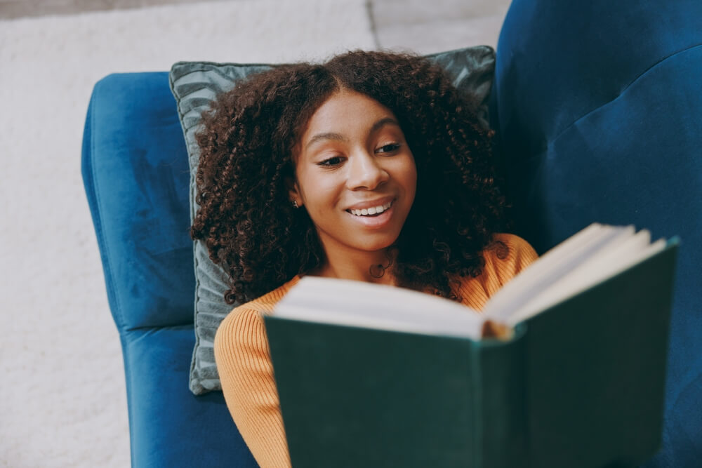 Imagem de uma mulher negra deitada em um sofá lendo um livro de capa verde. 