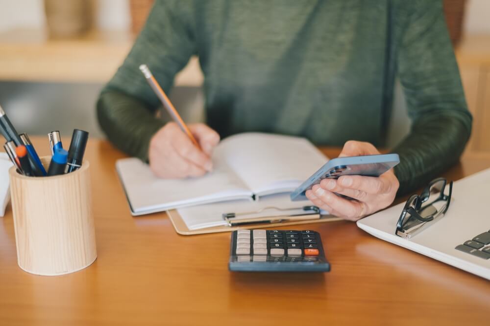 Imagem de um homem sentado em uma mesa fazendo anotações em um caderno ve mexendo no celular. Perto dele há uma calculadora.