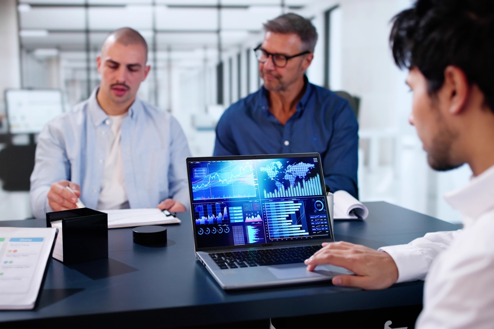 Imagem de três homens sentados em uma mesa. Um deles está mexendo em um computador, realizando uma análise preditiva.