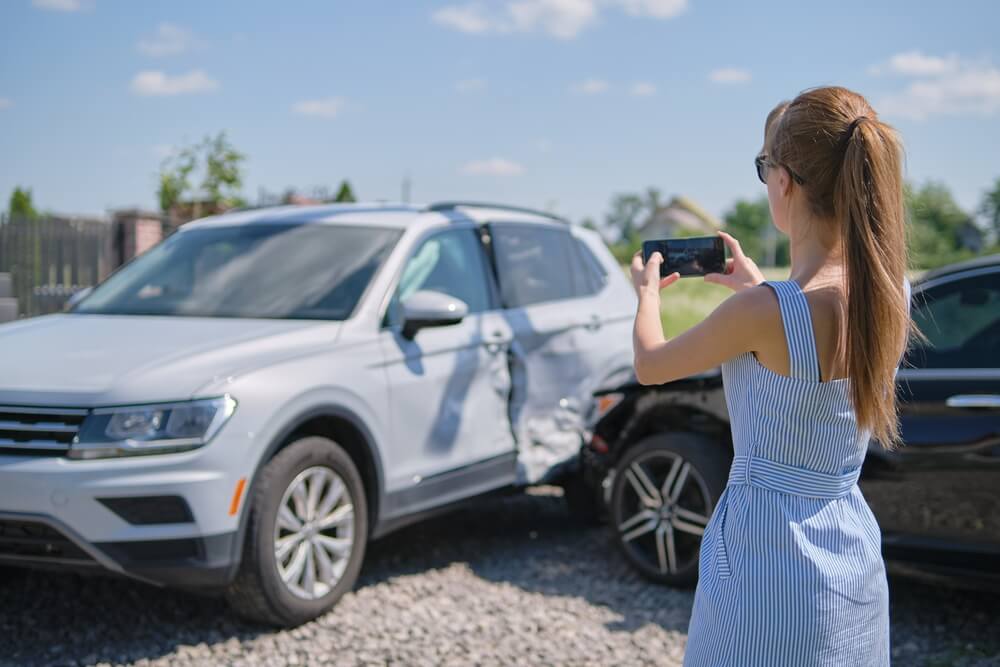 Imagem de uma mulher fotografando um carro após terem batido nele. A ideia é enviar a foto para o seguro.