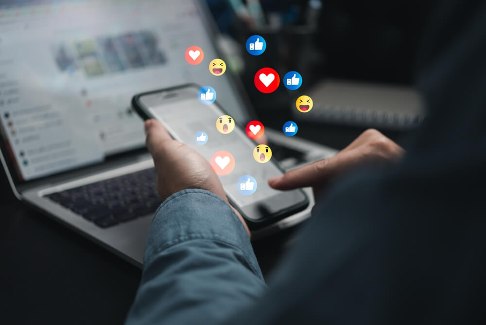Imagem de uma pessoa utilizando o celular em frente a um computador. Na tela do celular estão saindo figurinhas que representam reações disponíveis nas redes sociais.