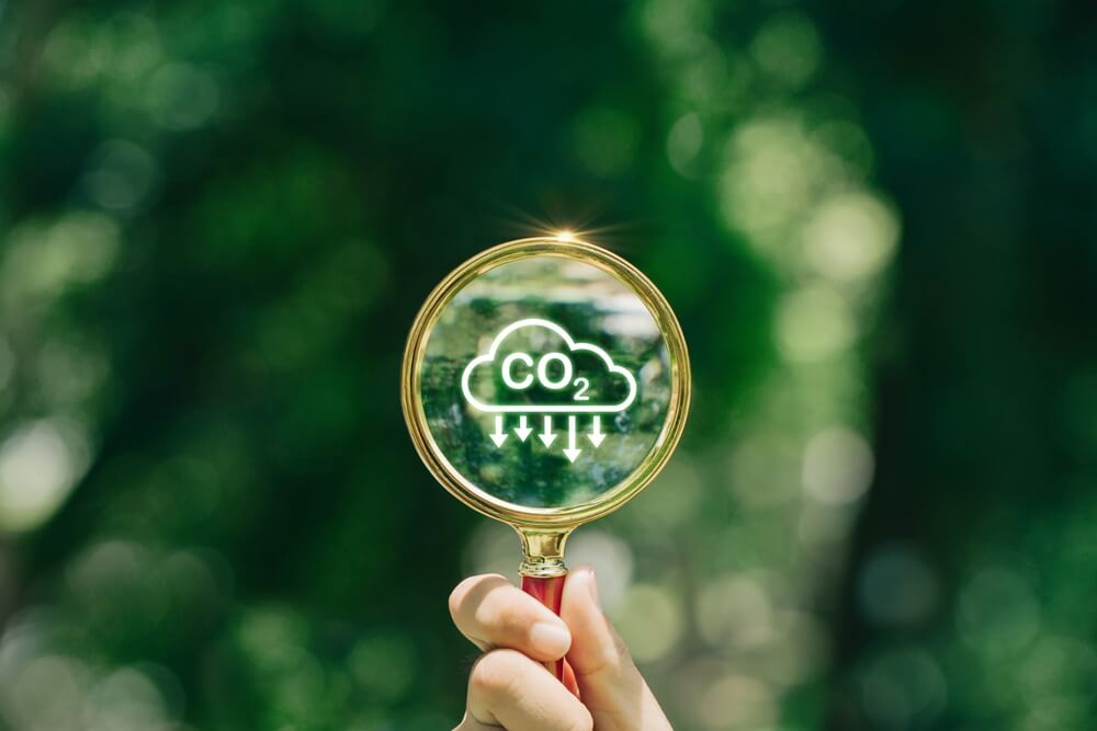 Imagem de uma lupa. Dentro dela é possível ver a imagem de um símbolo representando o CO². Ao fundo, é possível ver um fundo verde.