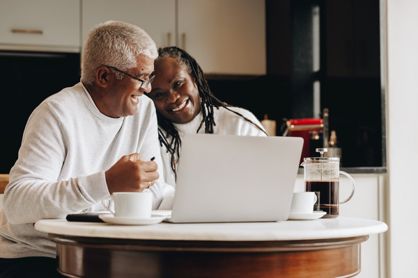 Imagem de um casal de aposentados, um homem e uma mulher, ambos negros, conferindo as finanças em um notebook. Eles estão felizes. Ao lado, há uma prensa de café.