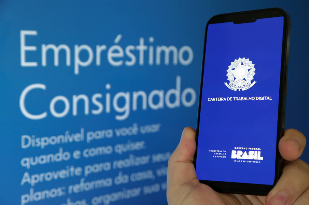 Imagem de um celular no primeiro plano com o app da Carteira de Trabalho iniciando. Ao fundo, é possível ler "Empréstimo Consignado".