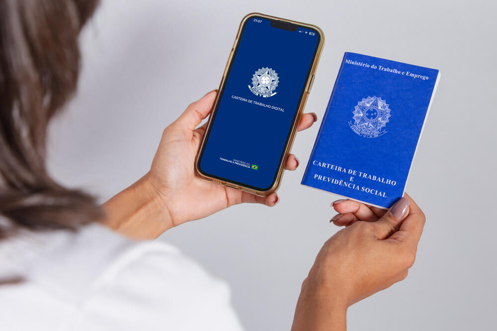 Imagem de uma mulher segurando um celular em uma mão, com a tela do aplicativo da carteira de trabalho aberta. Na outra mão, ela está segurando uma carteira de trabalho.