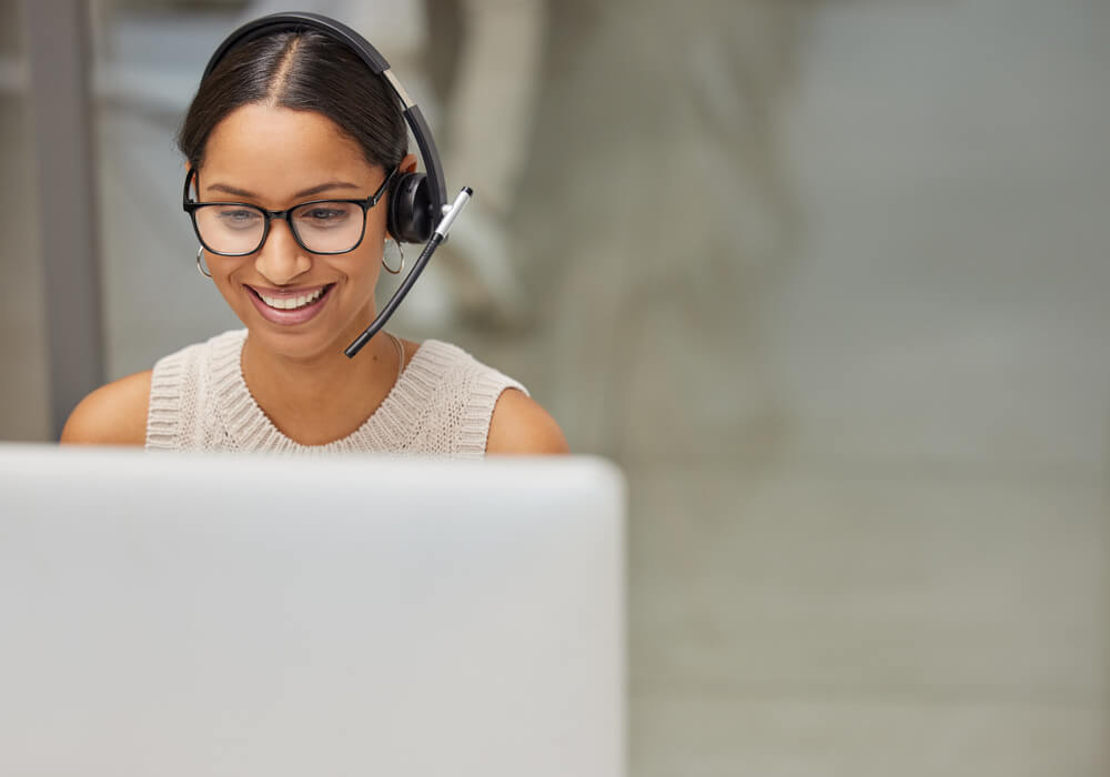 Imagem de uma mulher sentada em frente a um computador. Ela está mexendo no computador e com fones de ouvido, realizando um atendimento.