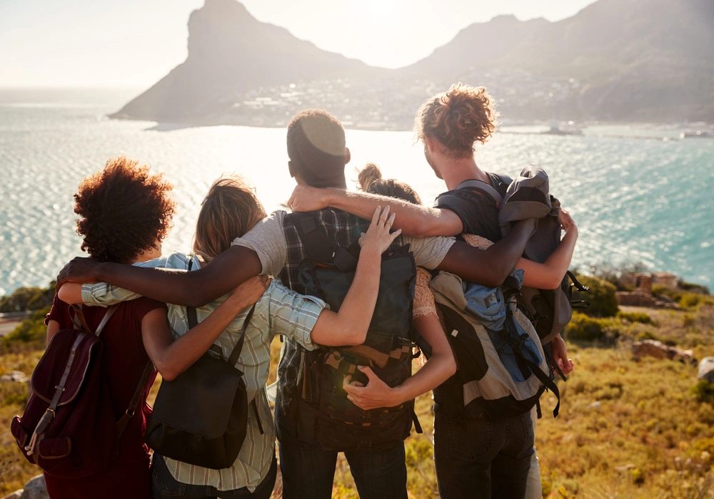 Imagem de jovens da geração Z viajando juntos. Eles estão de costas para a câmera, abraçados e admirando a paisagem.