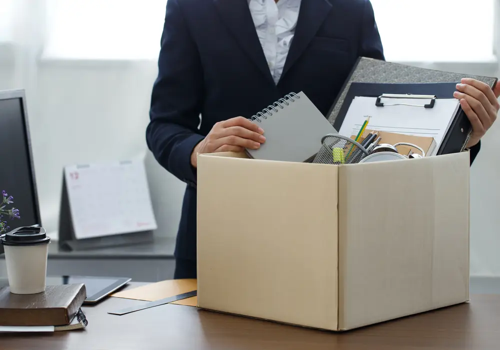 Imagem de uma pessoa, aparentemente uma mulher, colocando as coisas que estavam no escritório dele dentro de uma caixa de papelão, representando uma demissão.