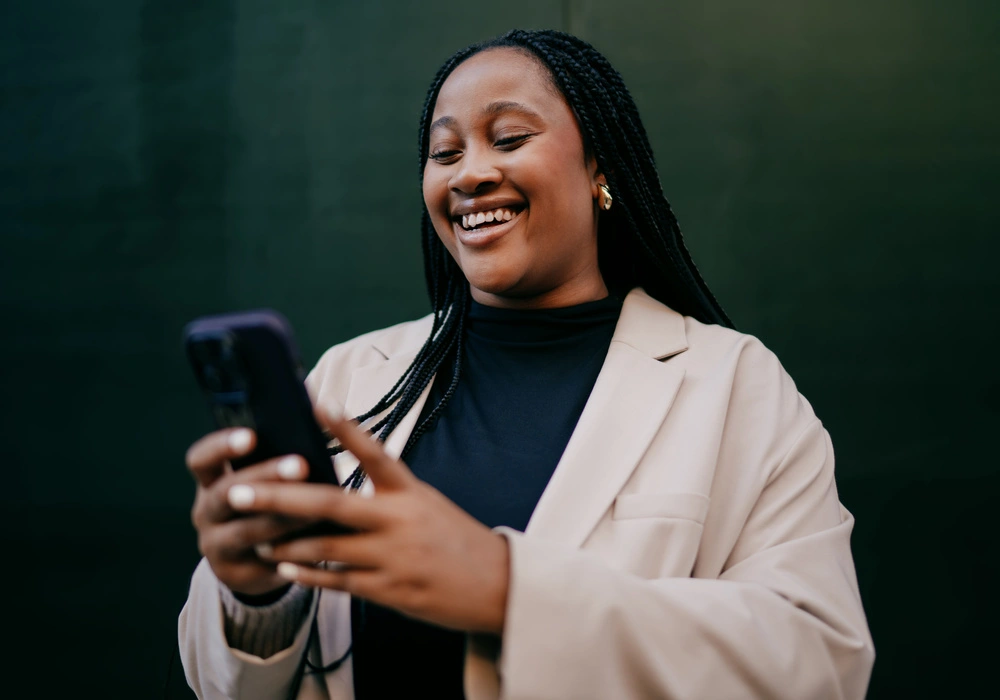 Imagem de uma mulher negra, jovem, feliz olhando o celular. Ela está contente, pois o saldo do Crédito do Trabalhador foi depositado na conta dela.